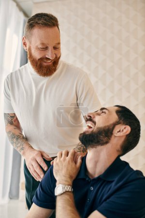 Two men, one with a beard, laughing together in a modern living room. Happy gay couple in casual clothes show love and joy.