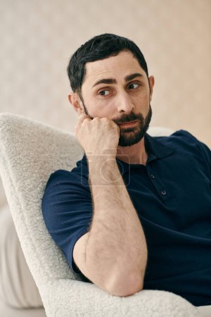 Photo for A man in casual clothes, sitting in a chair, deep in thought with his hand on his chin, in a modern living room. - Royalty Free Image