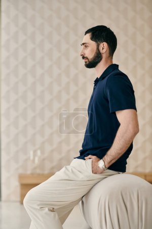 A man in casual clothes sits on a stool in a modern living room, lost in thought.