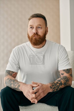 Téléchargez les photos : Homme barbu avec des tatouages s'assoit confortablement sur un canapé, respirant la confiance et l'expression de soi. - en image libre de droit