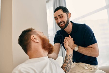 Foto de Two men in casual clothes hold hands lovingly in a modern living room, expressing happiness in their relationship. - Imagen libre de derechos