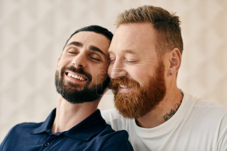 Two men with beards, one in a blue shirt and the other in a striped shirt, are smiling warmly at each other in a cozy living room.