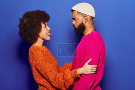 A young African American man and woman, friends, stand together in vibrant casual attire against a blue background, showing their bond.