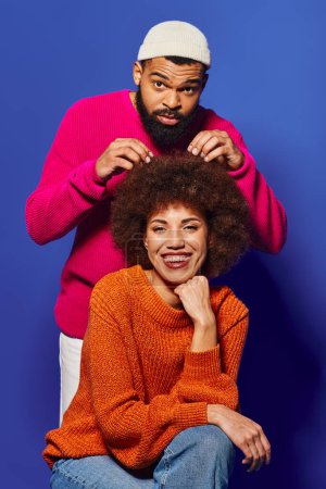 Foto de Un hombre y una mujer afroamericanos jóvenes se sientan juntos, mostrando un atuendo casual vibrante y un fuerte vínculo de amistad sobre un fondo azul. - Imagen libre de derechos