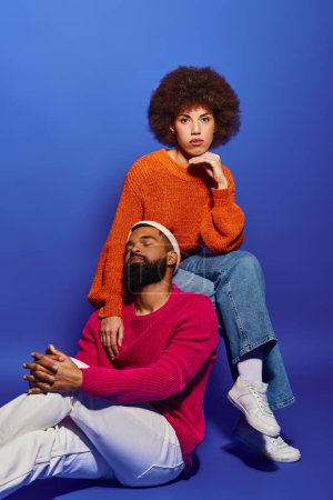 Photo for A young African American man and woman sit together, showcasing friendship and connection, against a vibrant blue backdrop. - Royalty Free Image