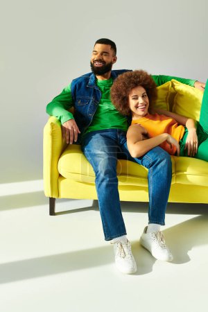 Foto de Felices amigos afroamericanos vestidos con ropa vibrante, un hombre y una mujer, sentados en un sofá amarillo sobre un fondo gris. - Imagen libre de derechos