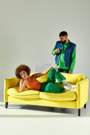 Ein Mann und eine Frau, fröhliche afroamerikanische Freunde in lebendiger Kleidung, sitzen auf einer gelben Couch vor grauem Hintergrund.