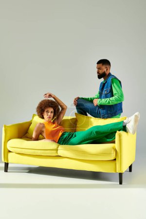 Glückliche afroamerikanische Freunde, Mann und Frau, sitzen auf einer gelben Couch in leuchtenden Kleidern und zeigen Freundschaft.