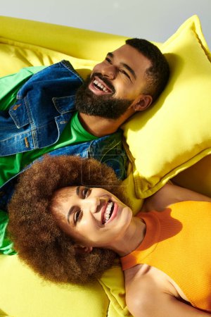 Ein afroamerikanischer Mann und eine afroamerikanische Frau in lebendiger Kleidung liegen friedlich auf einem gelben Kissen vor grauem Hintergrund..