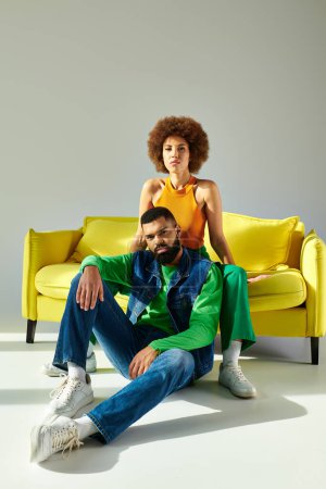 A happy African American man and woman in colorful outfits are sitting on a yellow couch against a grey background.