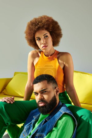Foto de Un hombre y una mujer afroamericanos vestidos de colores sentados felizmente en un sofá amarillo sobre un fondo gris. - Imagen libre de derechos