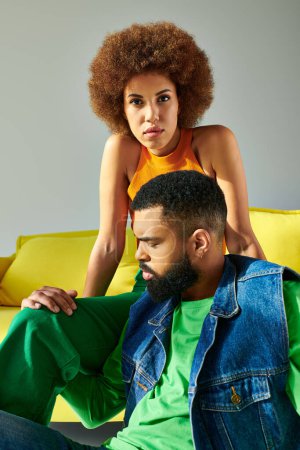 Téléchargez les photos : Un homme et une femme afro-américains en tenue colorée, s'appréciant mutuellement compagnie sur un canapé jaune vif sur fond gris. - en image libre de droit