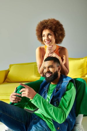 Photo for A happy African American man and woman sitting on a yellow couch, showcasing friendship in vibrant clothes on a grey background. - Royalty Free Image