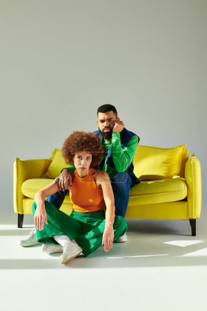 Foto de Amigos afroamericanos en traje colorido sentados en un sofá amarillo contra un fondo gris, mostrando amistad entre un hombre y una mujer. - Imagen libre de derechos