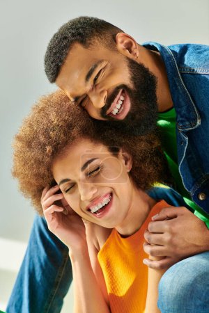 A happy African American couple in colorful attire smile warmly while embracing on a yellow couch against a grey background.