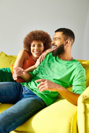 Un hombre y una mujer afroamericanos, vestidos vibrantemente, comparten un momento de amistad mientras están sentados en un sofá amarillo sobre un fondo gris.