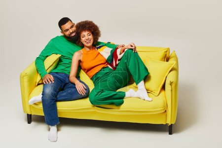 Happy African American friends in colorful attire sitting on a yellow couch against a grey backdrop, showcasing an intimate bond.