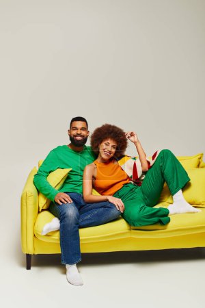 Téléchargez les photos : Heureux amis afro-américains en vêtements vibrants assis sur un canapé jaune sur un fond gris. - en image libre de droit