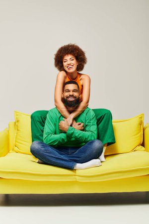 Fröhliche afroamerikanische Freunde in lebendiger Kleidung sitzen auf einer gelben Couch auf grauem Hintergrund und zeigen Freundschaft.
