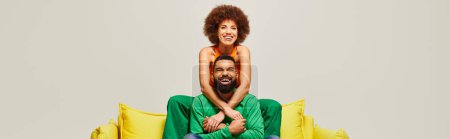 Téléchargez les photos : Un homme en tenue vibrante est assis au sommet d'une femme sur un canapé jaune, mettant en valeur le lien d'amitié dans un contraste coloré. - en image libre de droit