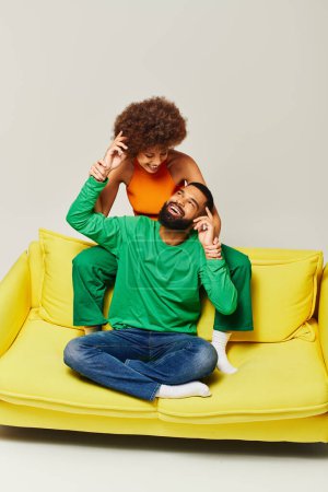 An African American man and woman happily sit on a yellow couch in vibrant clothes against a grey background.