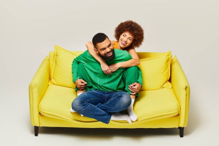 An African American man and woman, dressed in vibrant clothes, happily sit on a yellow couch against a grey background.