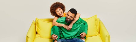 Photo for A happy African American man and woman, dressed in vibrant clothes, sit on a yellow chair against a grey background. - Royalty Free Image