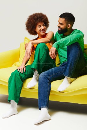 Happy African American man and woman in colorful attire sitting on yellow couch against grey backdrop.