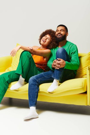 Foto de Un hombre y una mujer afroamericanos felices con ropa vibrante sentados en un sofá amarillo sobre un fondo gris. - Imagen libre de derechos