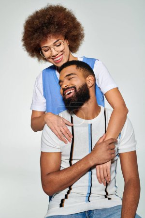 Amigos afroamericanos con ropa elegante muestran amistad mientras el hombre lleva a la mujer en su espalda contra un fondo gris.