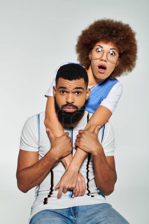 Ein afroamerikanischer Mann hebt und stützt stilvoll eine Frau auf seinen Schultern, während er vor grauem Hintergrund posiert.