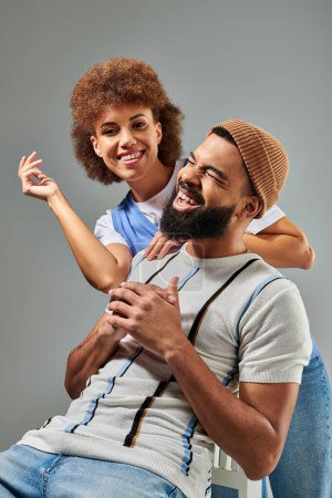 Téléchargez les photos : Un homme afro-américain élégamment habillé assis sur une chaise avec une femme sur le dos, affichant un moment d'amitié unique. - en image libre de droit