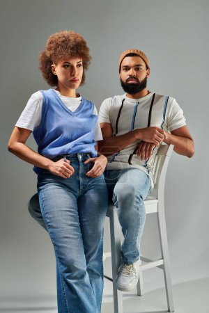Photo for A man and woman, friends, in stylish attire, sitting on a chair against a grey background, showcasing their friendship. - Royalty Free Image