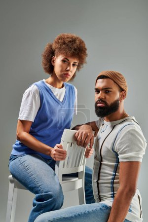 An African American man and woman in stylish clothes exhibit friendship while sitting on grey background.