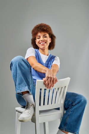 Téléchargez les photos : Une femme afro-américaine élégante en tenue chic s'assoit élégamment sur une chaise blanche sur un fond gris. - en image libre de droit