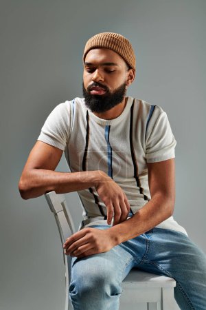 A stylishly dressed African American man with a beard sits elegantly on a chair against a grey background.