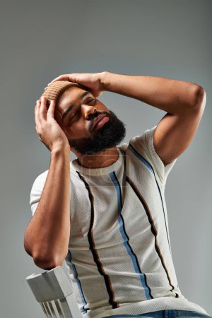 Téléchargez les photos : Un homme afro-américain élégamment habillé avec une barbe assise confortablement sur une chaise sur un fond gris. - en image libre de droit