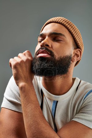 Photo for A man with a beard and a stylish hat poses confidently in fashionable attire on a grey background. - Royalty Free Image