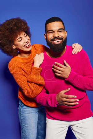 Un joven afroamericano hombre y mujer, amigos posando juguetonamente en vibrante atuendo casual sobre un fondo azul.