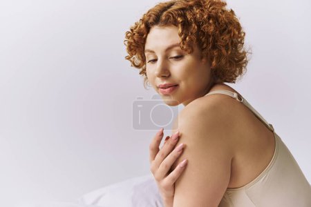 A young, curvy woman with fiery curls lounging on a cozy bed in lingerie against a serene grey backdrop.