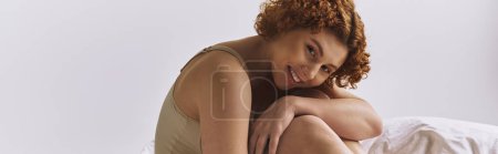 Téléchargez les photos : A young, curvy redhead woman in lingerie sitting with her legs crossed on a bed against a grey background. - en image libre de droit
