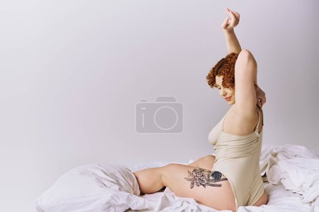 Foto de A young curvy redhead woman in lingerie sitting on a bed, raising her arms with a serene expression. - Imagen libre de derechos
