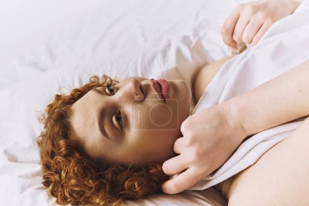 A young, curvy redhead woman in lingerie relaxing on a bed with a grey background.