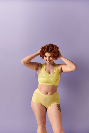 A young curvy redhead woman in a yellow bikini striking a pose against a bold purple backdrop.