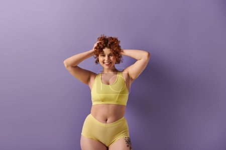 Téléchargez les photos : A young, curvy redhead woman confidently poses in a yellow bikini against a striking purple background. - en image libre de droit