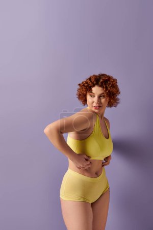 Curvy redhead woman in yellow bikini poses confidently in front of a vibrant purple wall.