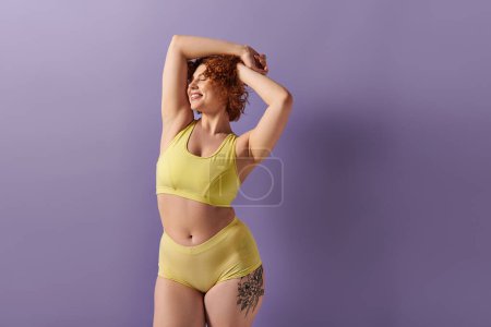 A young, curvy redhead woman in a yellow bikini striking a pose on a purple background.