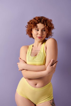 Young, curvy redhead woman in yellow bikini striking a confident pose against a vibrant purple backdrop.