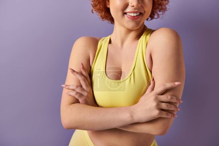 A young woman with fiery red hair strikes a confident pose in a vibrant yellow bikini against a bold purple backdrop.