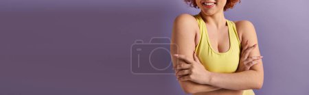 A young, curvy woman with vibrant red hair strikes a confident pose, crossing her arms, against a purple background.